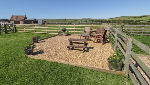 A fenced outdoor seating area with wooden benches and chairs, surrounded by gravel and grass. - Street House Farm Cottage (Saltburn-by-the-Sea)