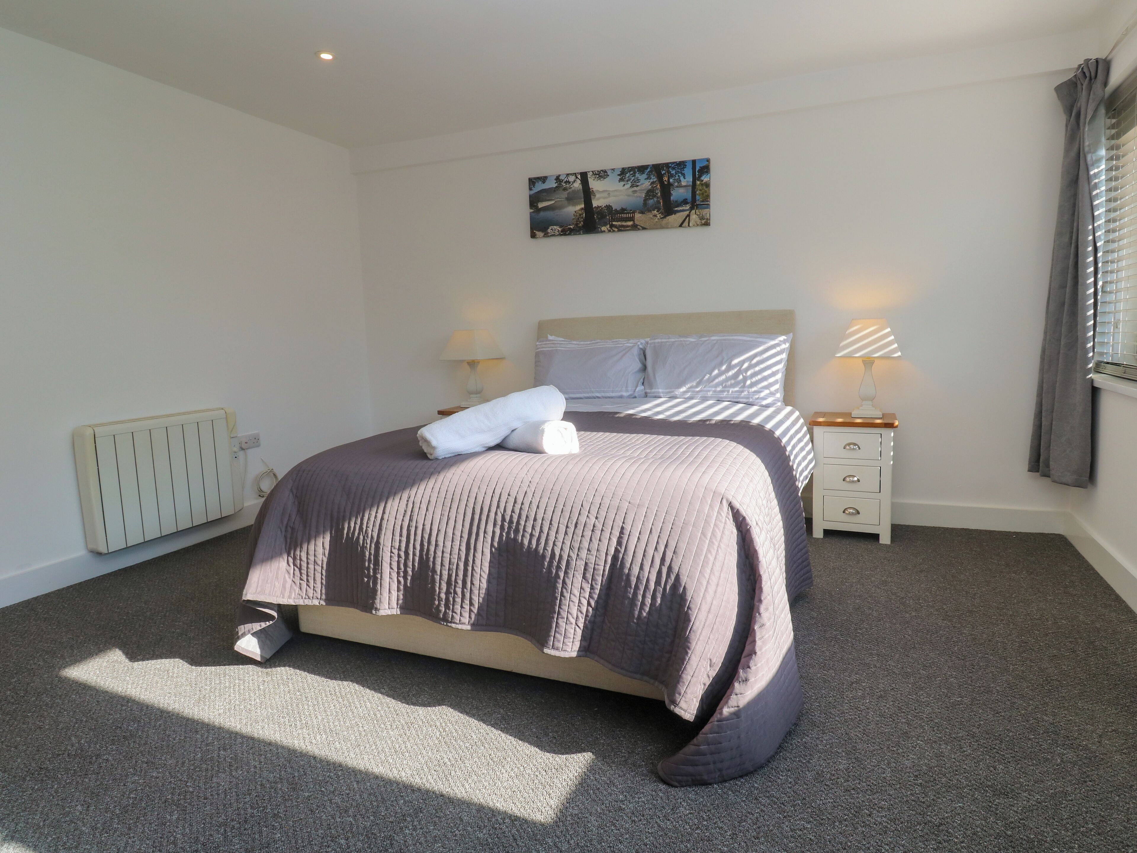 A neatly made bed with a headboard, two bedside tables with lamps, a radiator, and a painting on the wall.