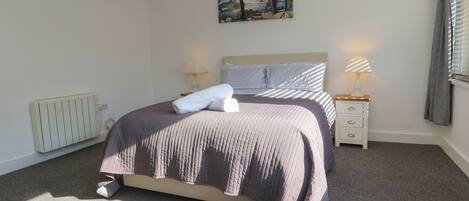 A neatly made bed with a headboard, two bedside tables with lamps, a radiator, and a painting on the wall.
