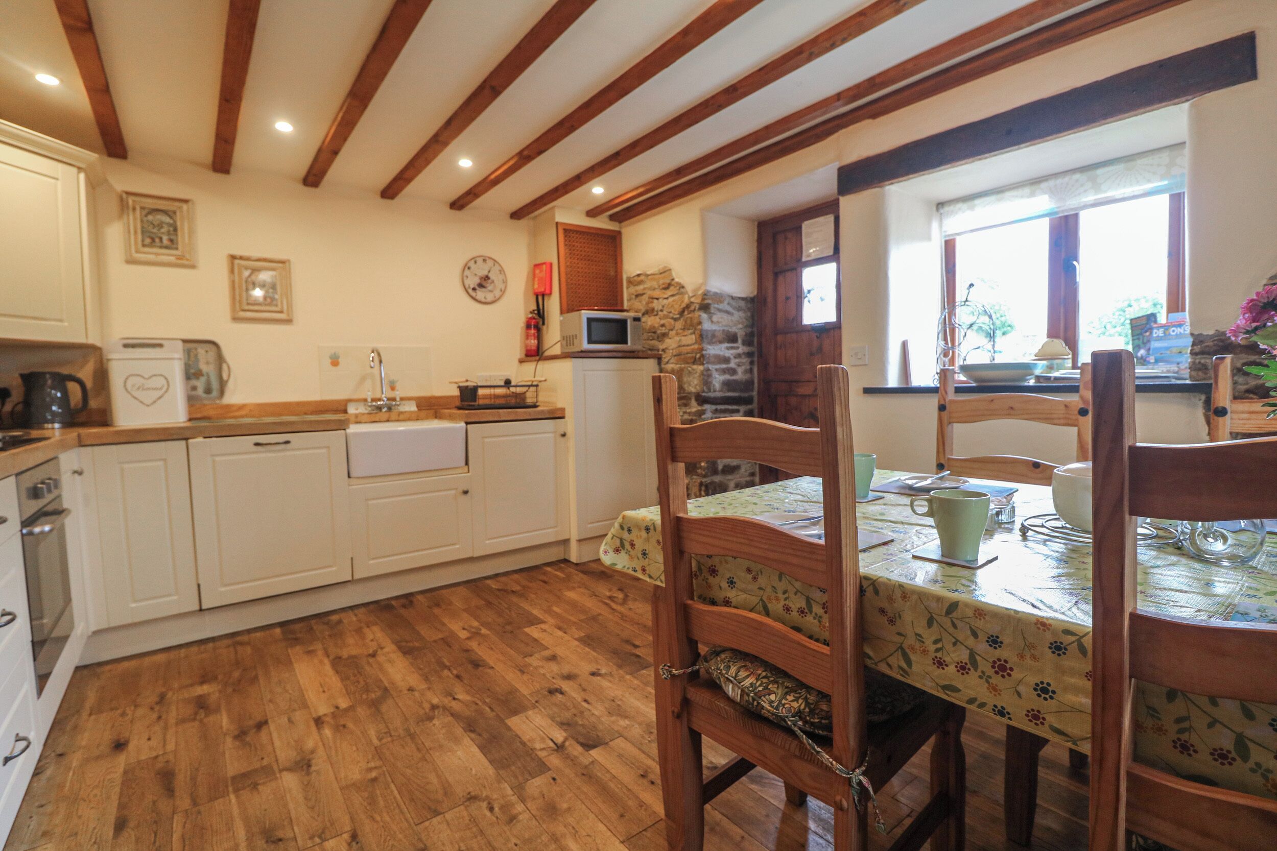 A kitchen with wooden beams, white cabinets, a wooden dining table with chairs, and a window with a view of greenery.