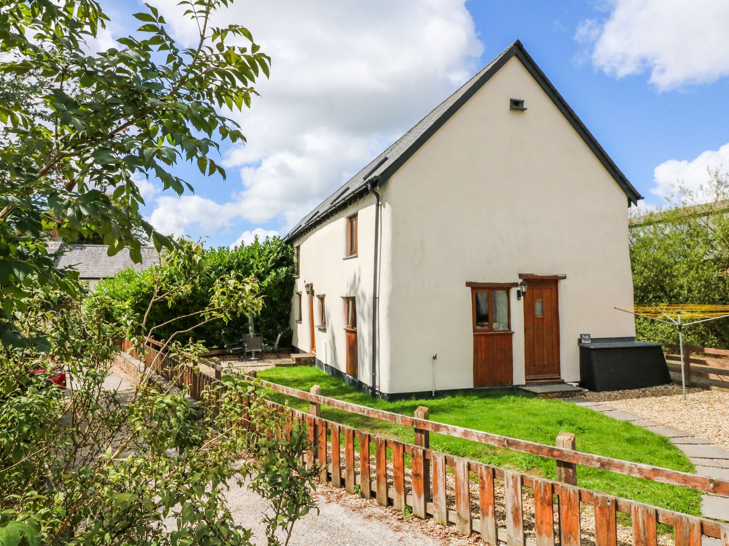 A white two-story house with a wooden door and windows, surrounded by a wooden fence and greenery.