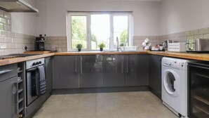 A modern kitchen with grey cabinets, a built-in oven, a washing machine, and a window providing natural light. - Palm Court (Carnforth)