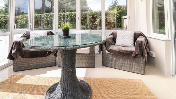 A conservatory with a glass table surrounded by wicker furniture, a potted plant, and a view of greenery outside. - Palm Court (Carnforth)