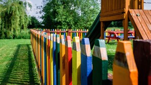 A wooden play structure with a slide and climbing area, surrounded by a fence with colorful painted posts.