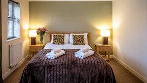 A neatly made bed with a brown striped bedspread, two pillows with leaf patterns, and two rolled towels. There are two bedside lamps, a window with blinds, and a radiator on the wall.
