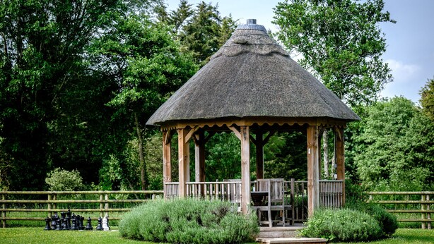 A gazebo with a thatched roof, surrounded by a well-maintained lawn and a wooden fence.