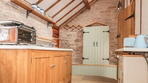 A rustic kitchen with wooden cabinets, a stone wall, and a lofted ceiling. - The Cow Cott (Hereford)