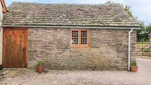 A stone building with a wooden door and window, potted plants, and a concrete ground. - The Cow Cott (Hereford)