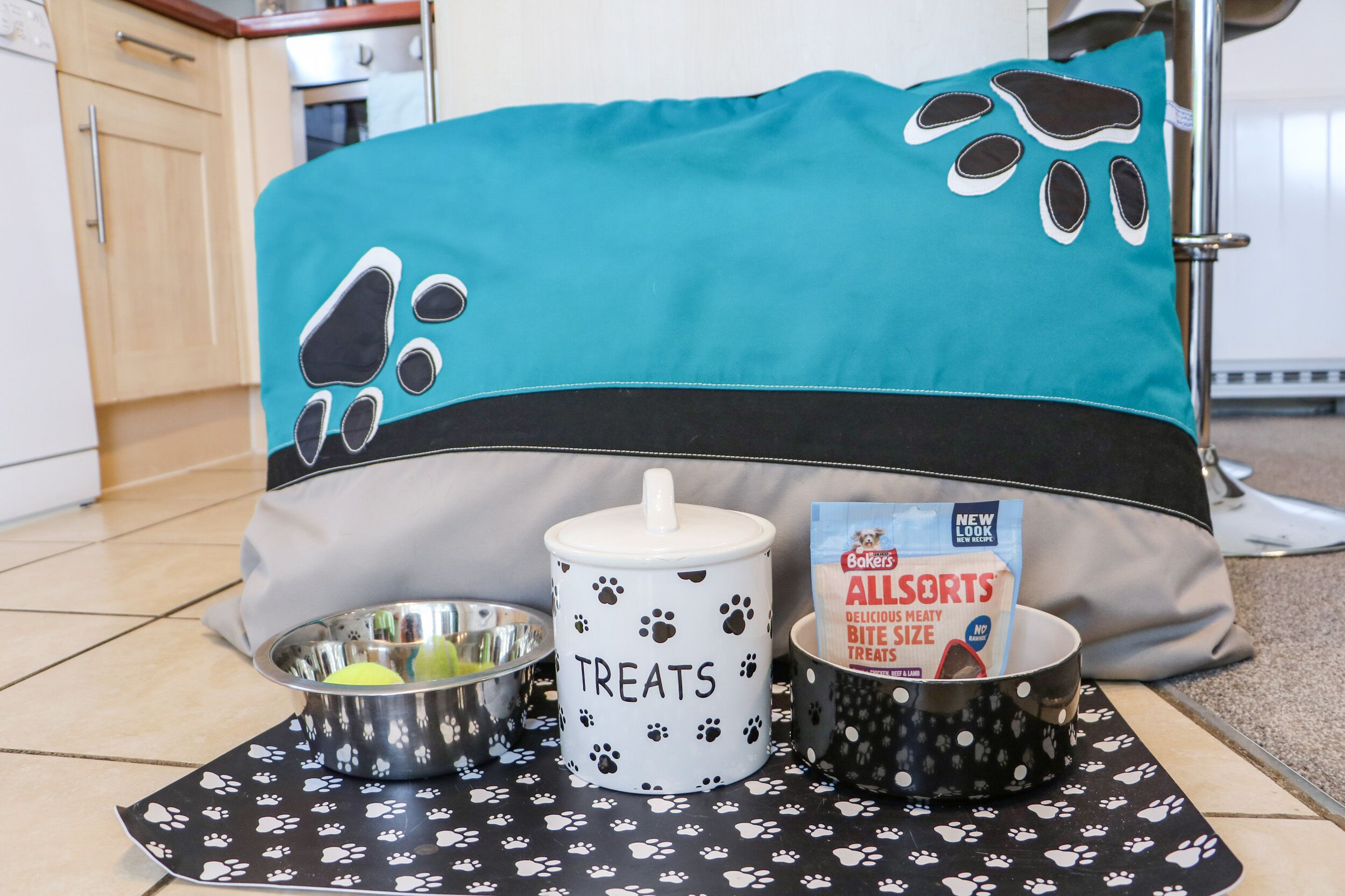 Una cama para mascotas con diseño de huella negra y verde azulado, un cuenco de golosinas y un bote de premios All's Meaty tamaño.
