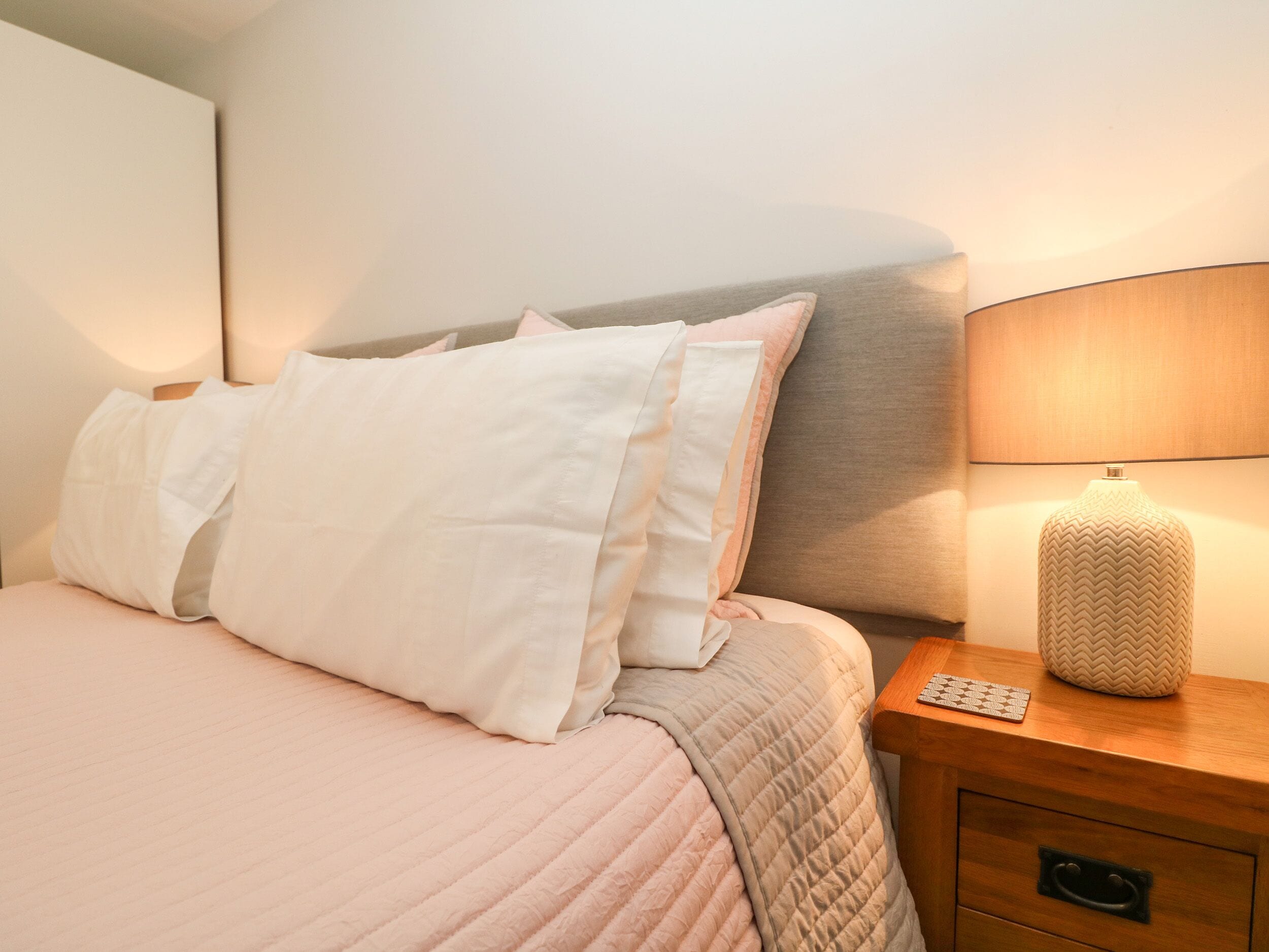 A neatly made bed with a pink quilt and white pillows, a wooden nightstand with a lamp, and a small drawer.