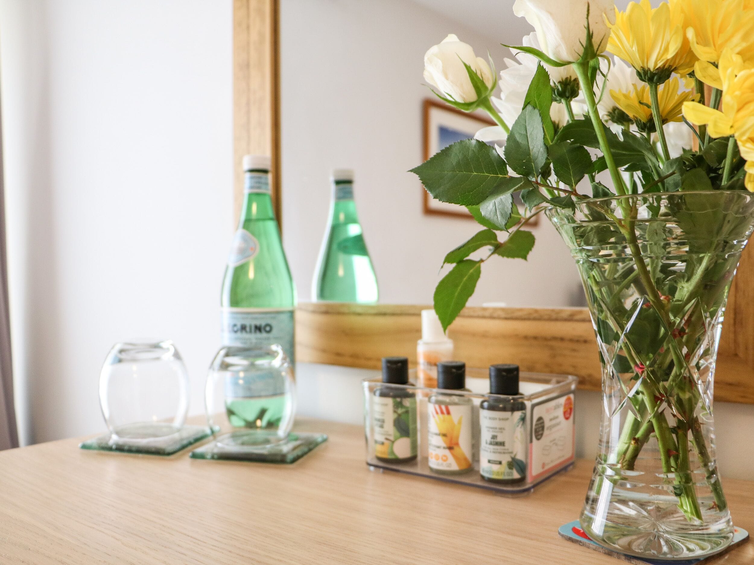 Een glazen vaas gevuld met witte en gele bloemen op een houten tafel, samen met een paar groene flessen en een klein flesje lotion.