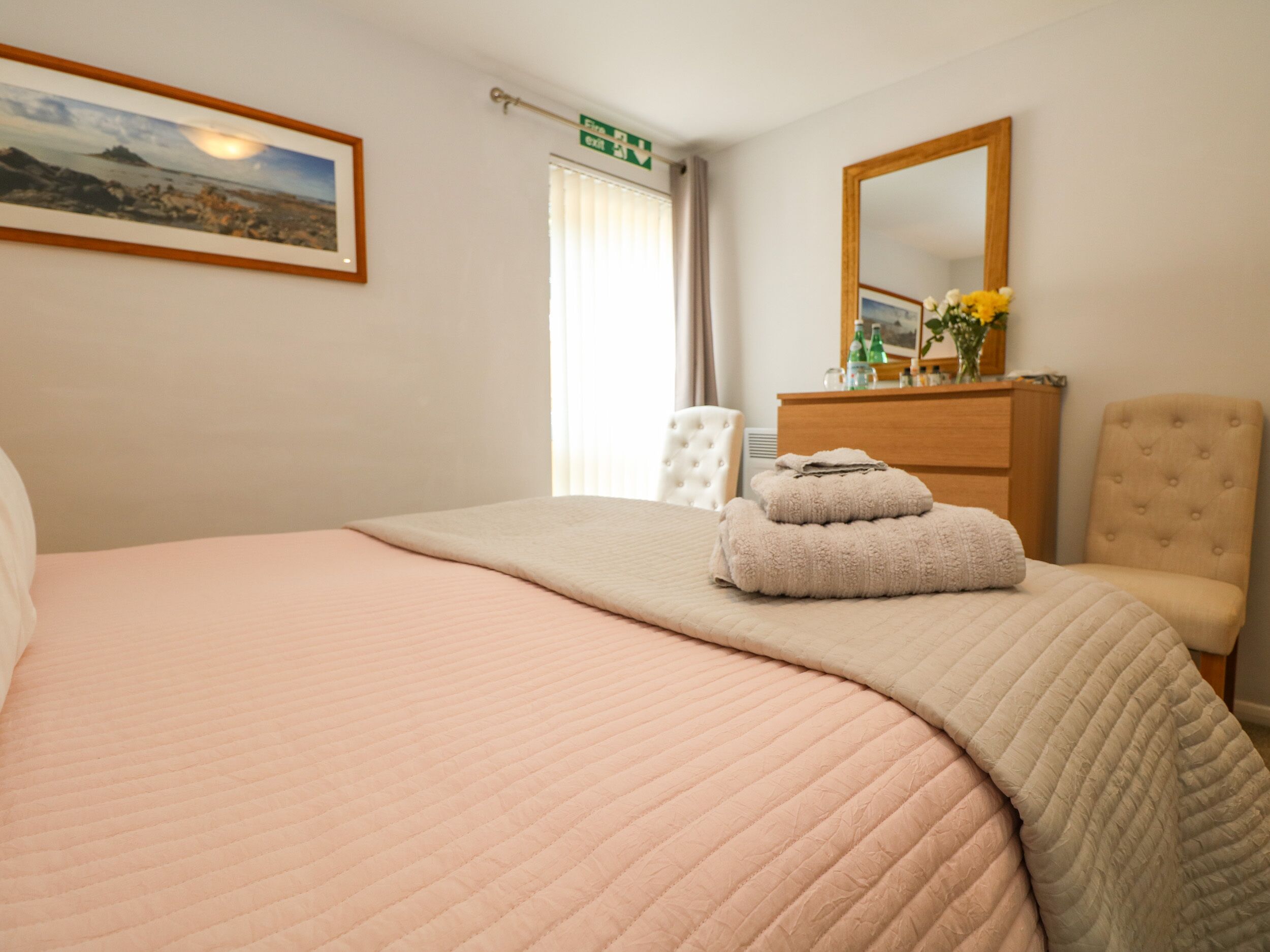 Una cama bien tendida con colcha rosa, un aparador de madera con artículos de tocador, un espejo y un cuadro en la pared.