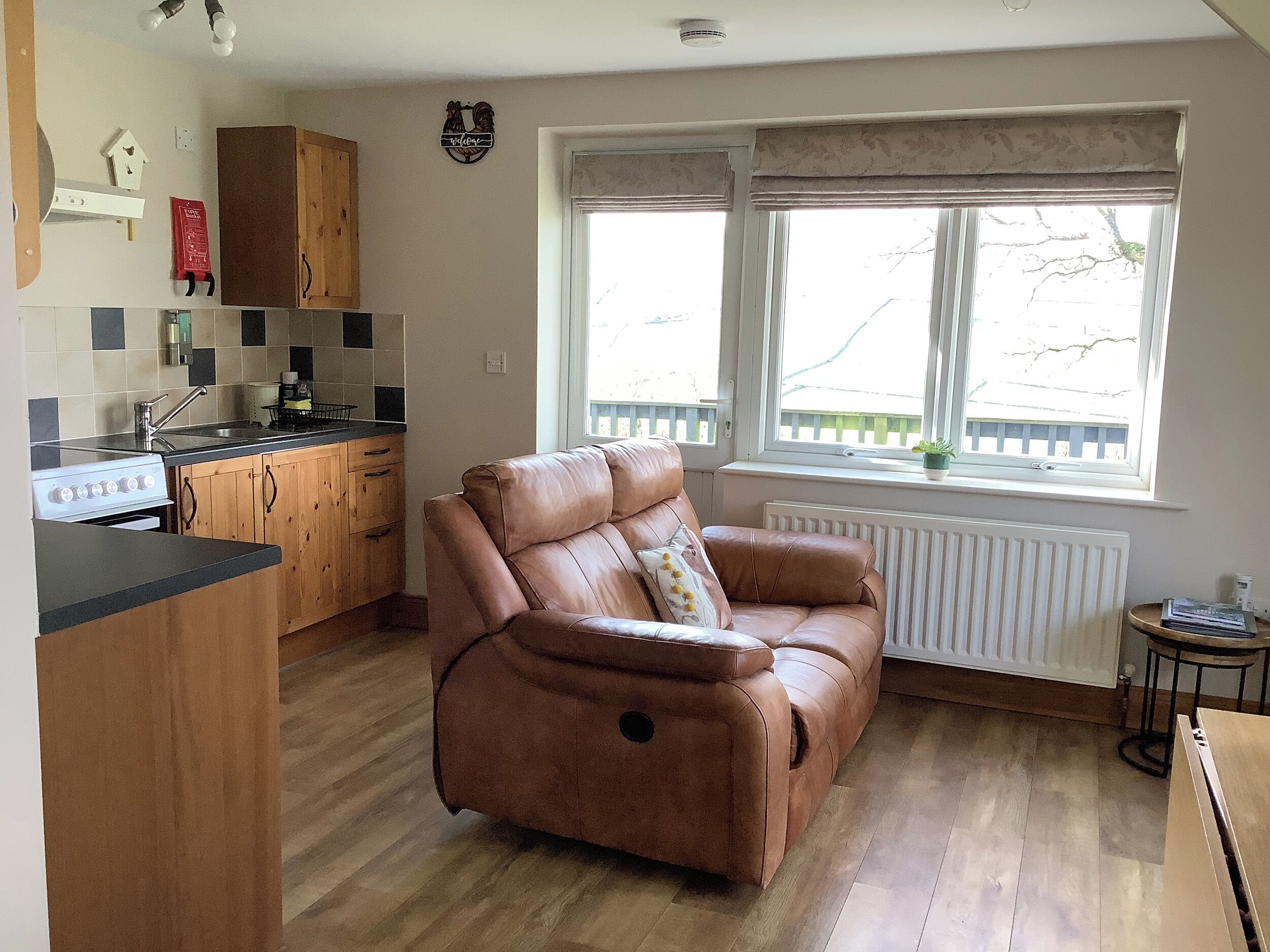 A cozy living room with a brown leather sofa, a kitchen area with wooden cabinets, and a large window providing natural light.