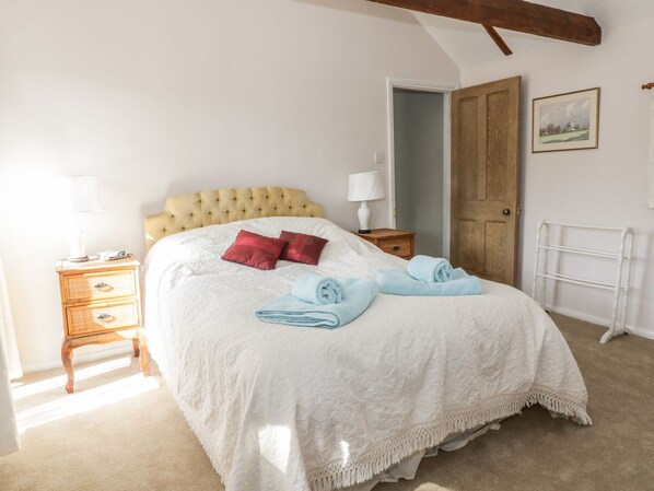 A neatly made bed with a white bedspread and two red pillows, a wooden nightstand with a lamp, and a white radiator. - Stable Cottage (Sudbury)