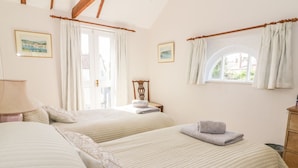 A bedroom with two beds, a wooden headboard, a window with curtains, and a small table with a lamp. - Stable Cottage (Sudbury)