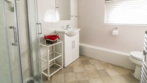 A bathroom with a glass shower enclosure, a white sink, a mirror above the sink, a red basket on a white shelf, a toilet, and a window with blinds. - Stable Cottage (Sudbury)