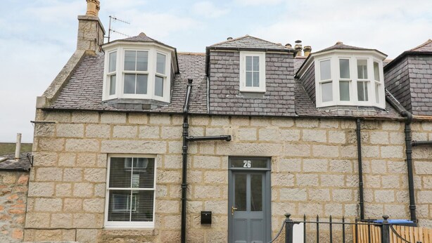A stone house with a dark grey door, two bay windows, and a number '26' on the wall. - Westwood (Ballater)