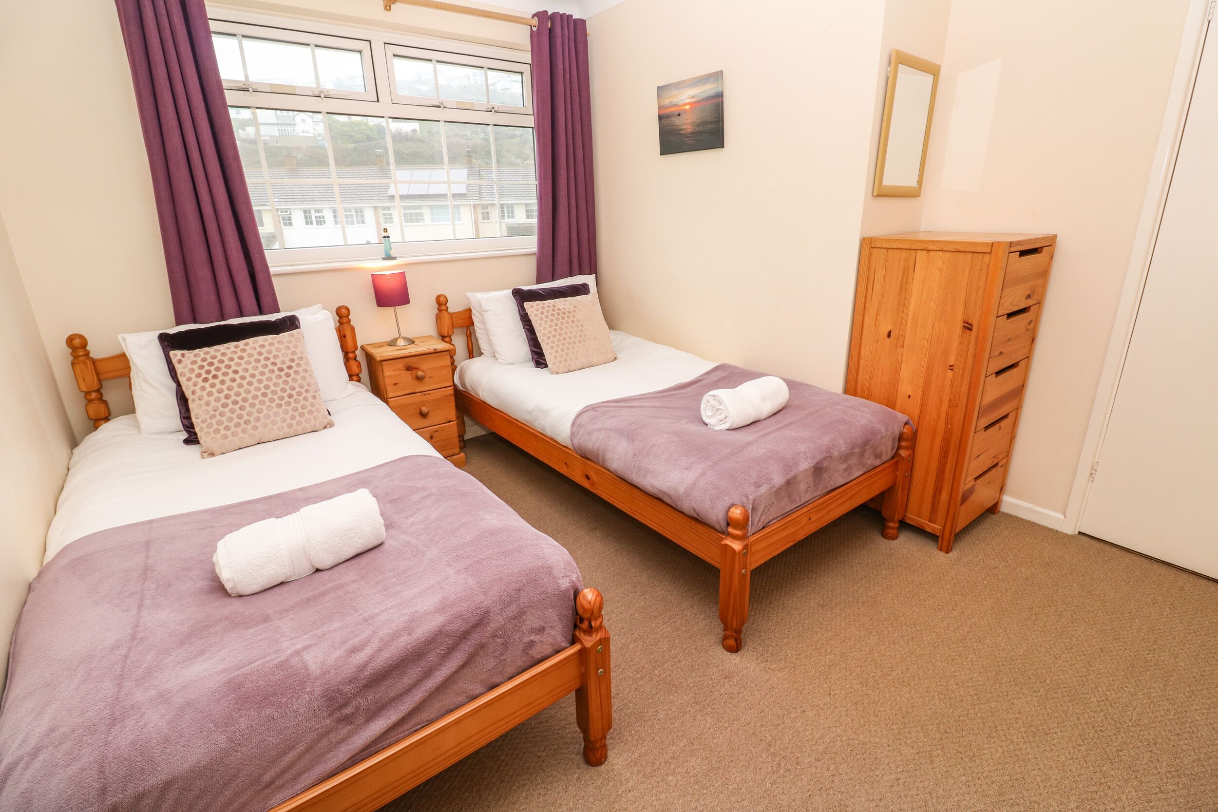 Quarto com duas camas de madeira, um guarda-roupa de madeira, uma mesa de cabeceira com abajur e uma janela com cortinas roxas.