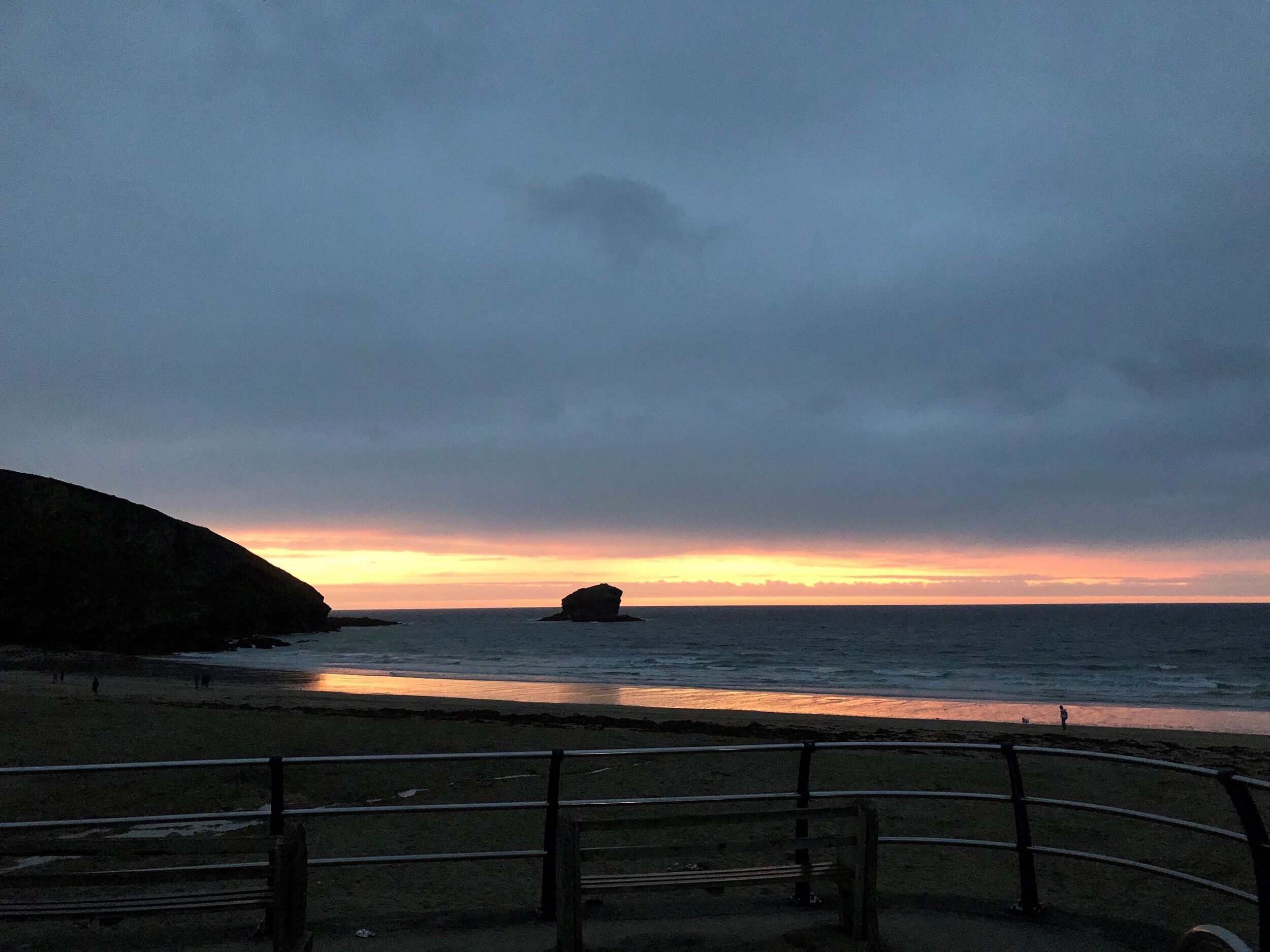 Ein Strand mit Geländer, ruhigem Wasser und einem Sonnenuntergang.