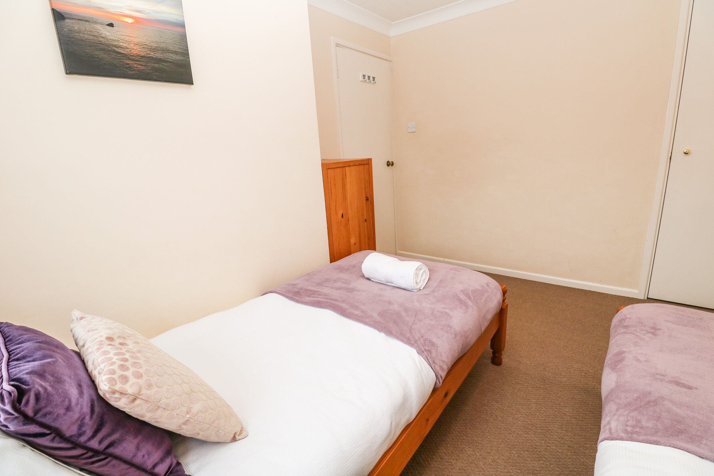 Una habitación con dos camas individuales, cada una con una manta morada y un almohadón blanco. Se ve una puerta de madera y una pared color beige.