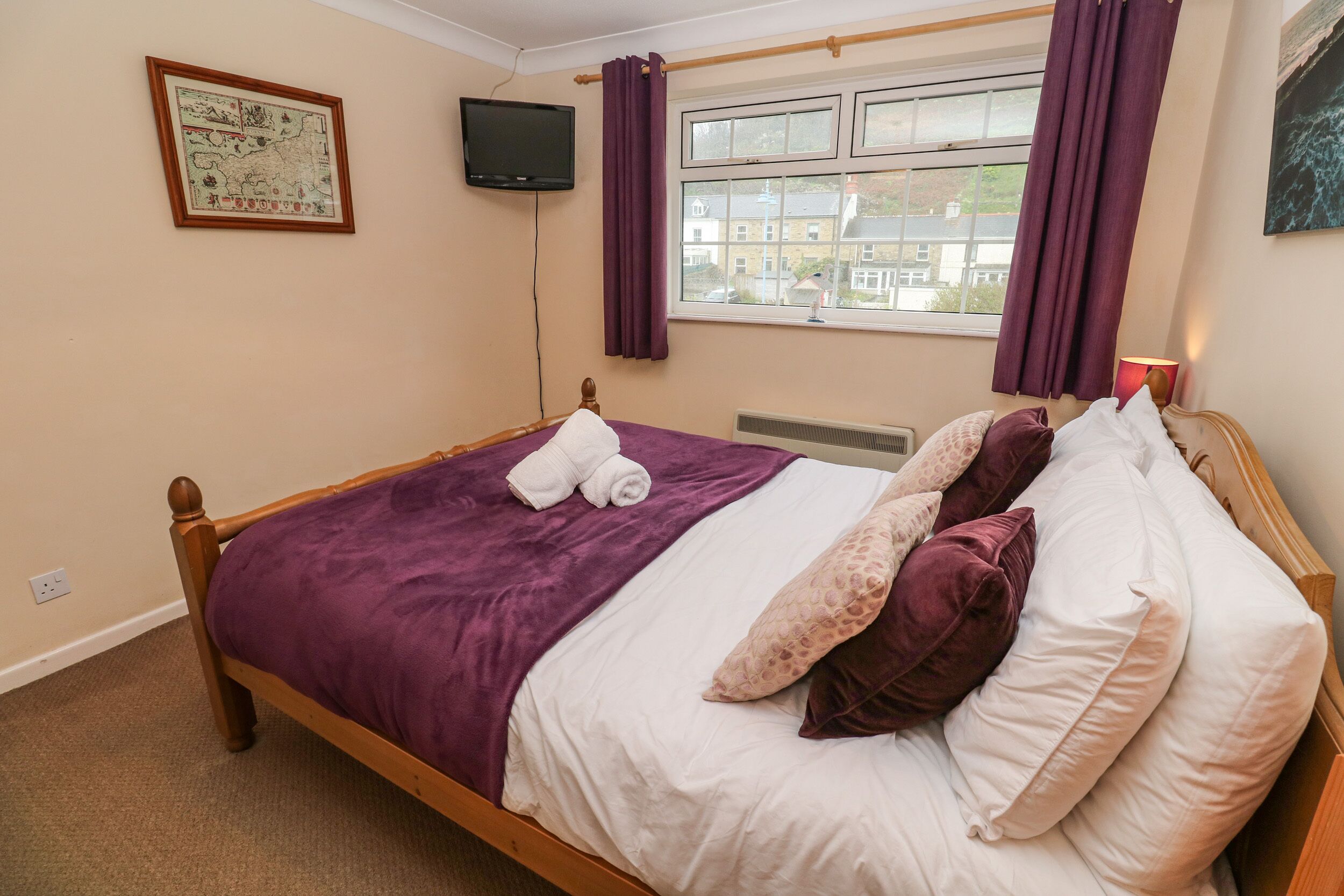 A neatly made bed with a purple blanket and white linens, a framed map on the wall, and a window with purple curtains.