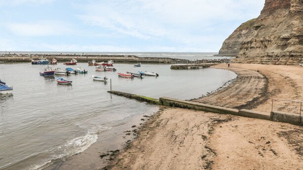 A coastal scene with boats docked along a sandy beach and a rocky cliff in the background. - Clevelyn (Saltburn-by-the-Sea)