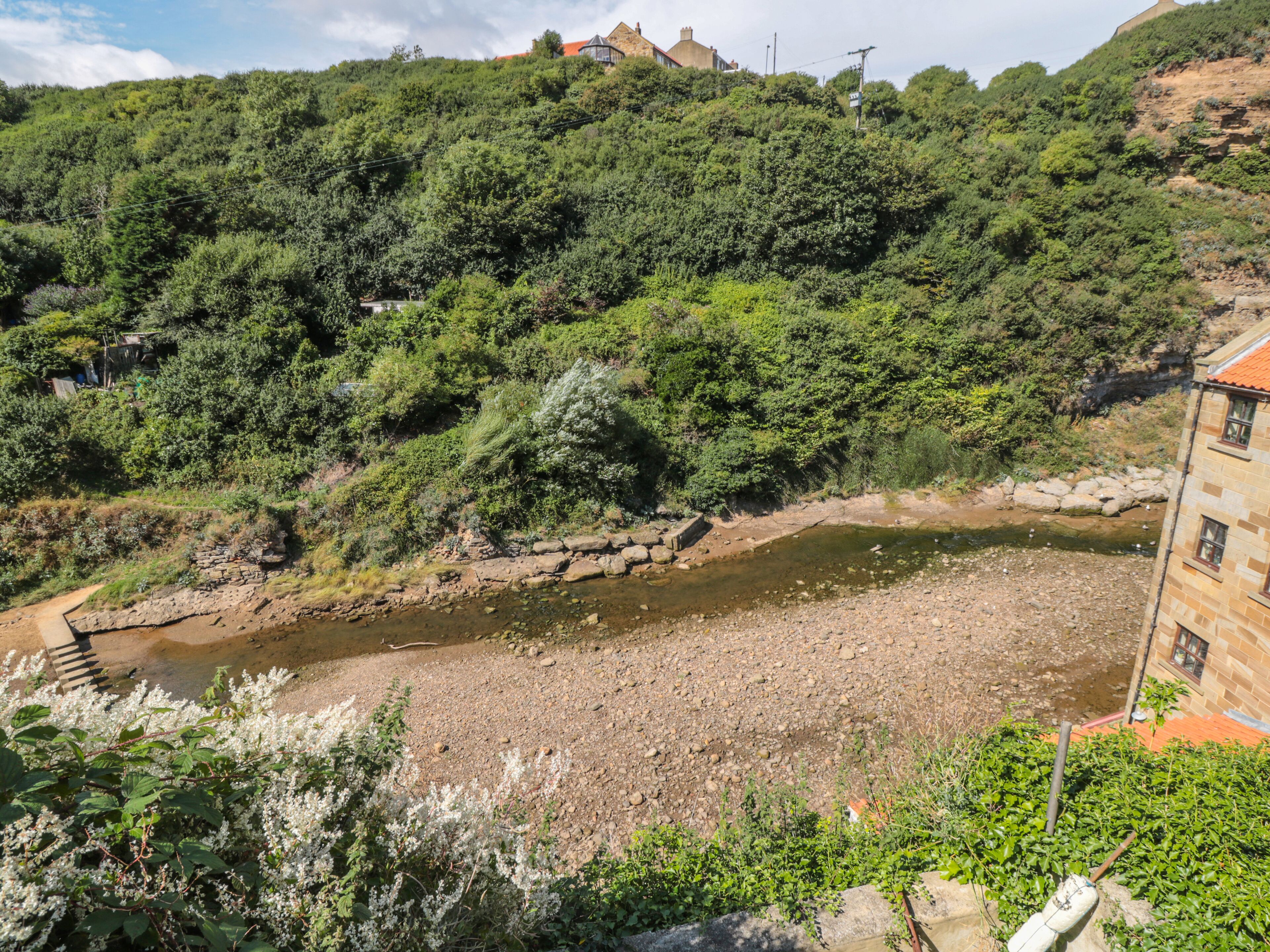 A riverside view with a stone bridge, lush greenery, and a building with a red roof.