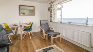 Ein Zimmer mit Esstisch, bunten Stühlen, einem grauen Sessel, einem Fenster mit Meerblick und einem weißen Heizkörper.