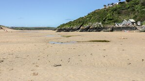 Ein sandiger Strand mit vereinzelten Pfützen, ein grasbewachsener Hügel mit Gebäuden und ein klarer Himmel.