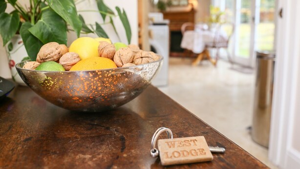 En skål med frukt och nötter står på ett träbord, och bredvid ligger en nyckelring med texten ”WEST LODGE”.