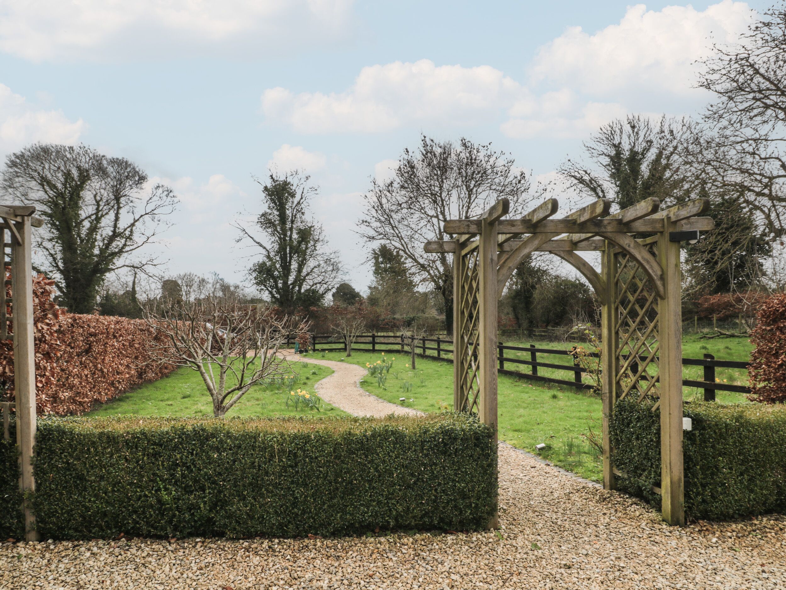 A garden pathway leading through a wooden pergola structure surrounded by hedges and trees.