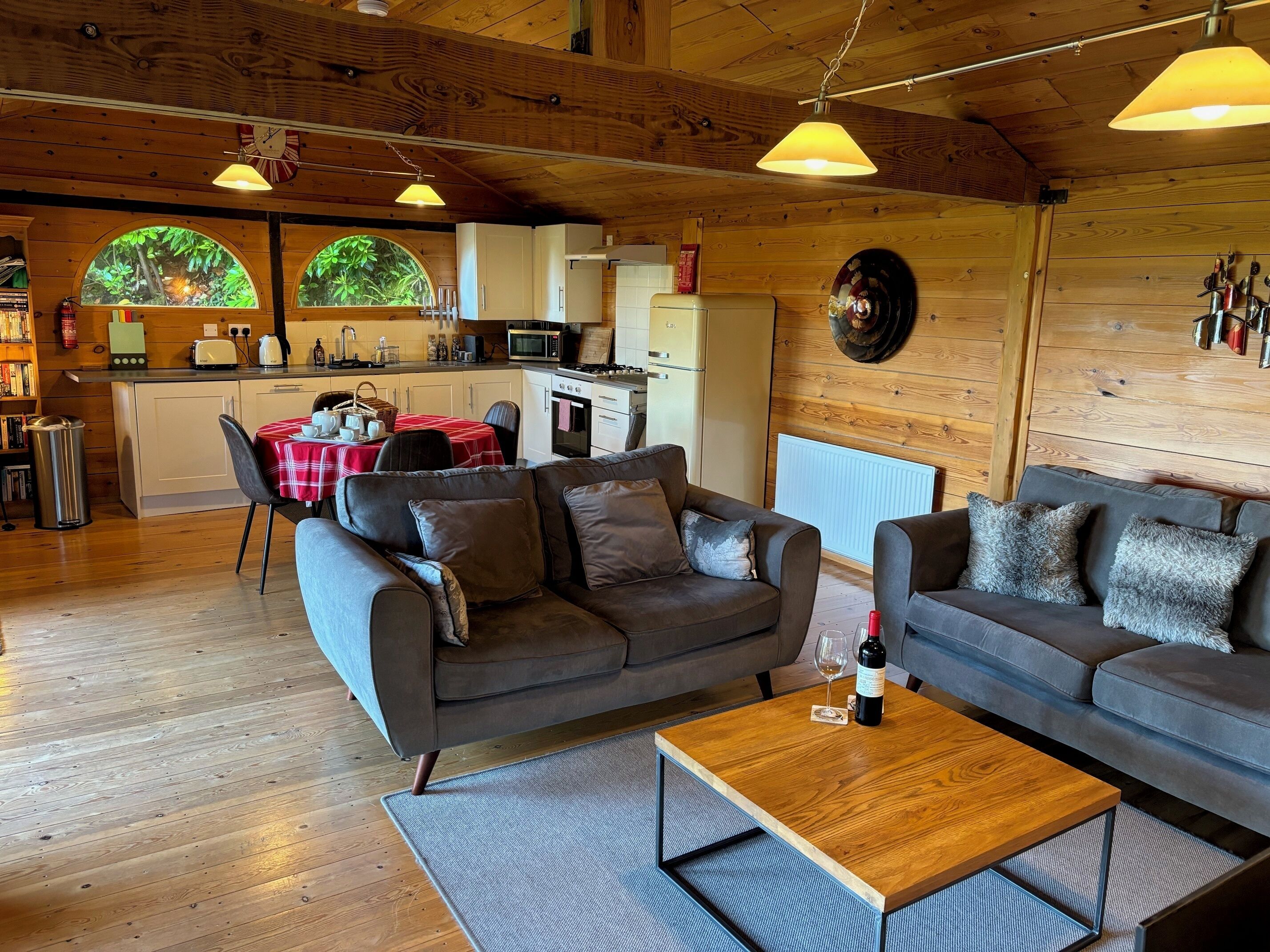 Ein gemütliches Blockhaus-Interieur mit Holzboden, zwei Sofas, einem Esstisch und einer Küchenzeile mit Blick auf viel Grün durch große Fenster.