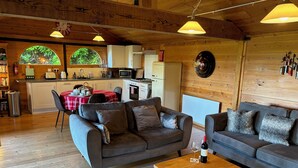 A cozy cabin interior with a wooden floor, two sofas, a dining table, and a kitchen area with a view of greenery through large windows. - Treetops (Barnstaple)