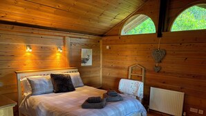 A wooden cabin bedroom with a bed, two wall-mounted lights, a window, and a radiator. - Treetops (Barnstaple)