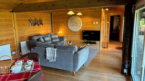 A cozy cabin interior with a grey sofa, wooden floors, a dining table with a red tablecloth, and a TV mounted on the wall. - Treetops (Barnstaple)