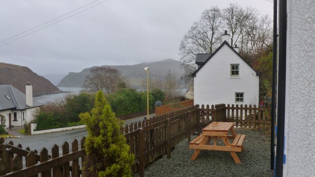 A white house with a wooden fence, a gravel patio, and a view of the sea and hills. - Sidhean Dubha Holiday Home (Portree)