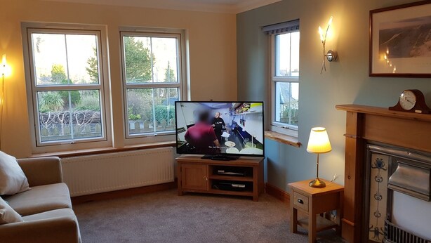 A living room with a television, a fireplace, and large windows. - Sidhean Dubha Holiday Home (Portree)