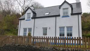 A two-story house with a stone wall in front, a wooden fence, and a sloped roof. - Sidhean Dubha Holiday Home (Portree)