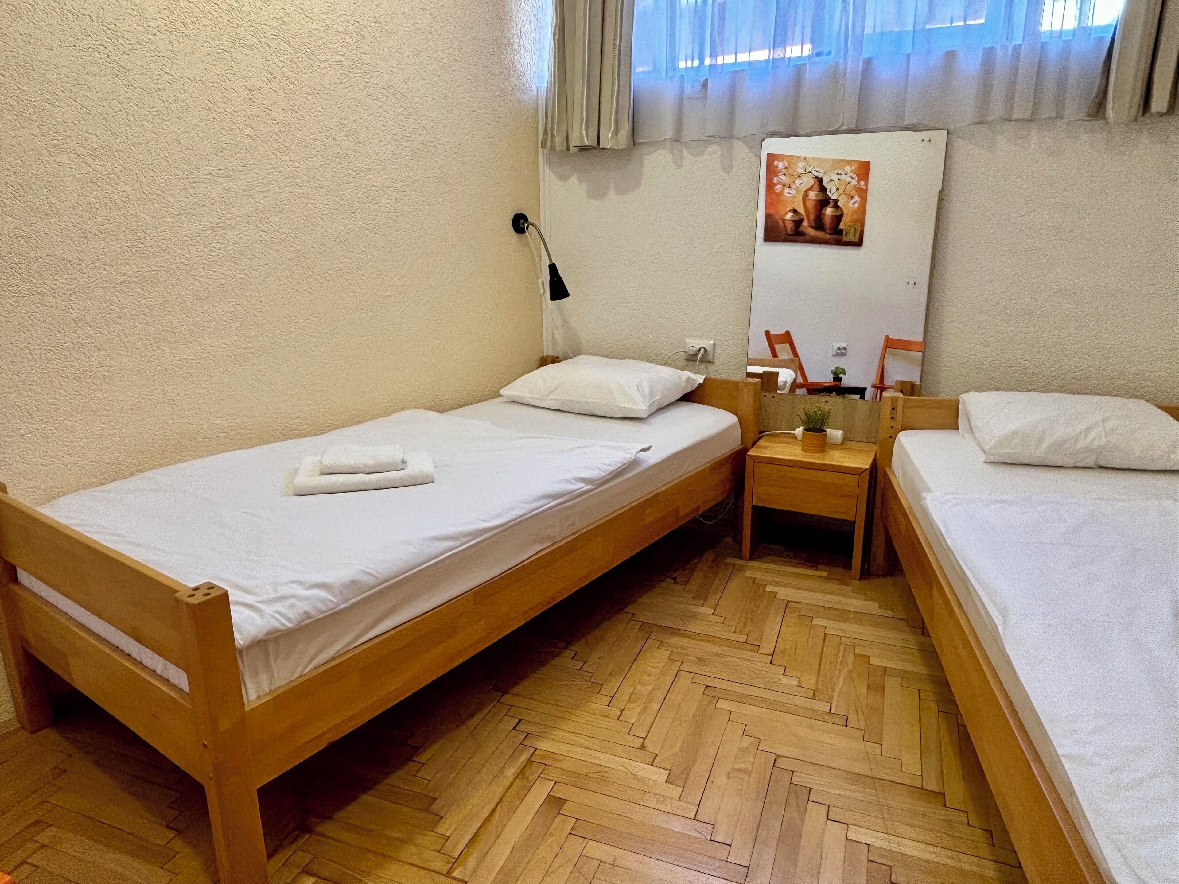 Une petite chambre propre et bien rangée, avec deux lits en bois, une table de chevet en bois sur laquelle se trouve une lampe, et un miroir accroché au mur.