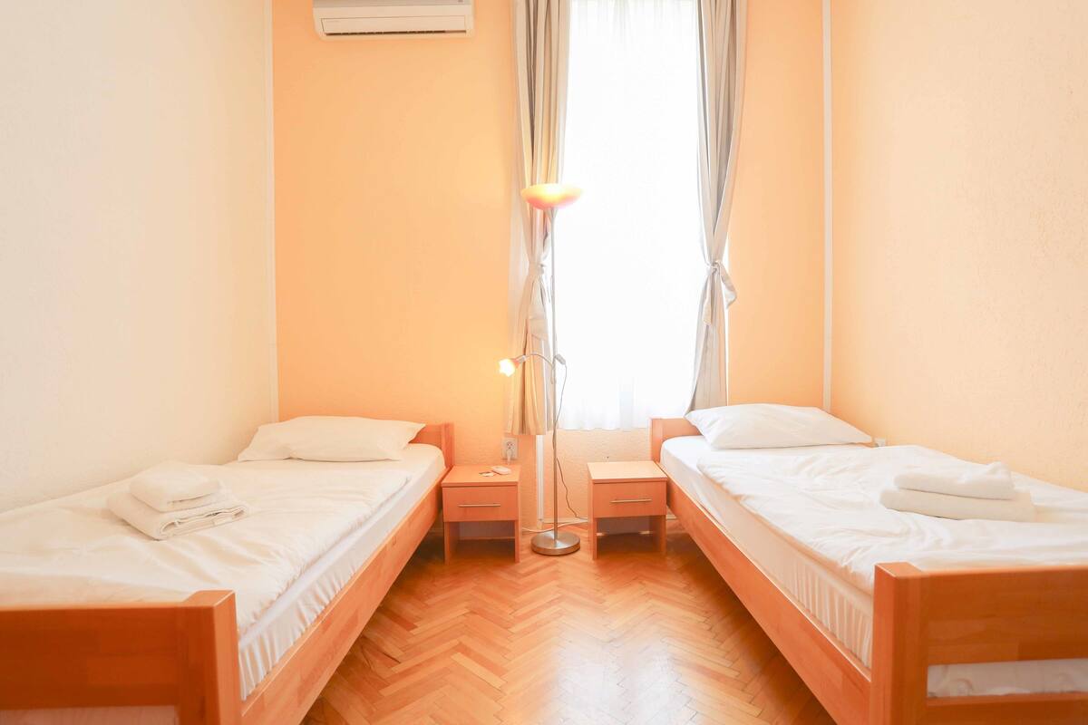 Una habitación con dos camas individuales, mesitas de noche de madera, una lámpara de pie y una ventana con cortinas.