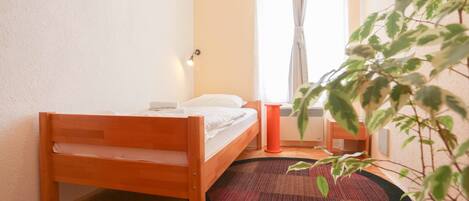 A small, neatly arranged bedroom with a wooden bed, a round rug, a potted plant, and a window with curtains.