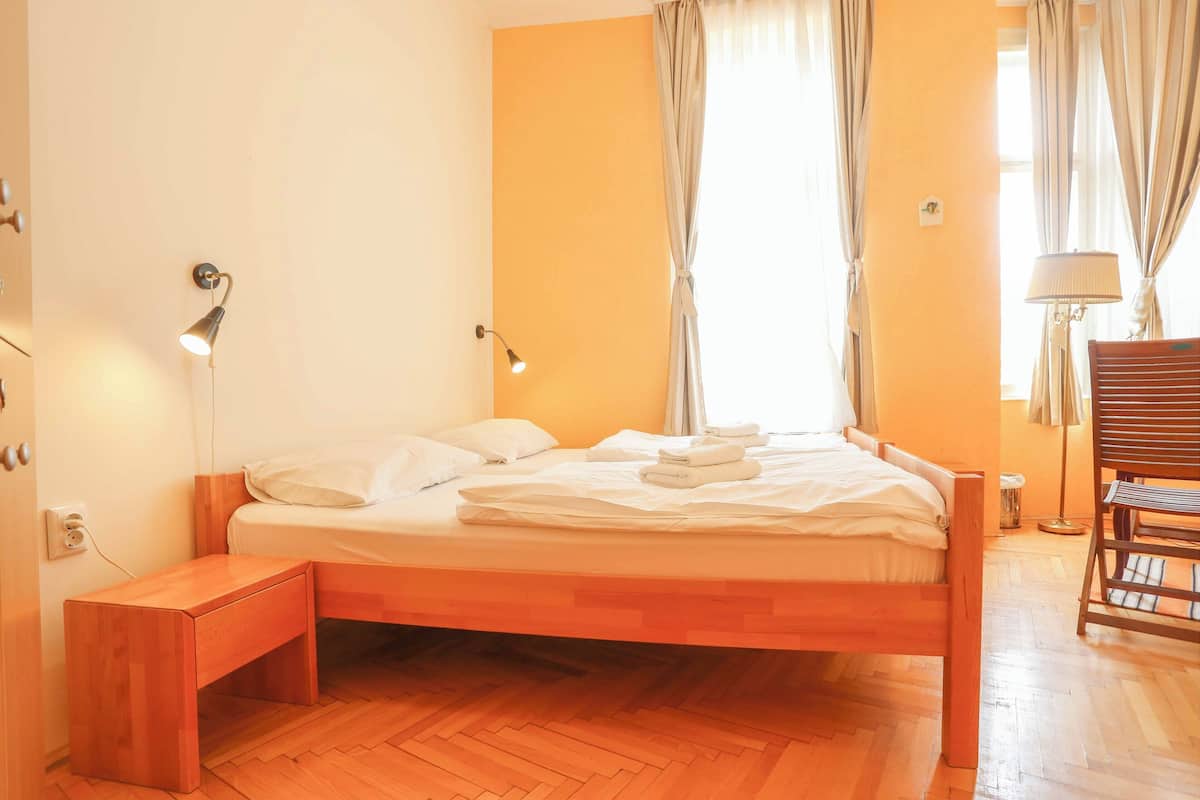 Una cama bien tendida con sábanas blancas, una mesita de noche de madera y una silla en una habitación con suelo de madera y una ventana con cortinas.