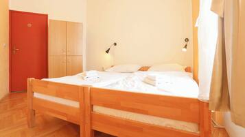 A double bed with white linens, a wooden wardrobe, a red door, and a wall-mounted lamp.
