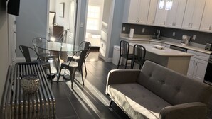 A modern kitchen with white cabinets, a dining area with a glass table and metal chairs, and a grey sofa. - Giles Ave 6 Bedroom Gem (Jersey City)