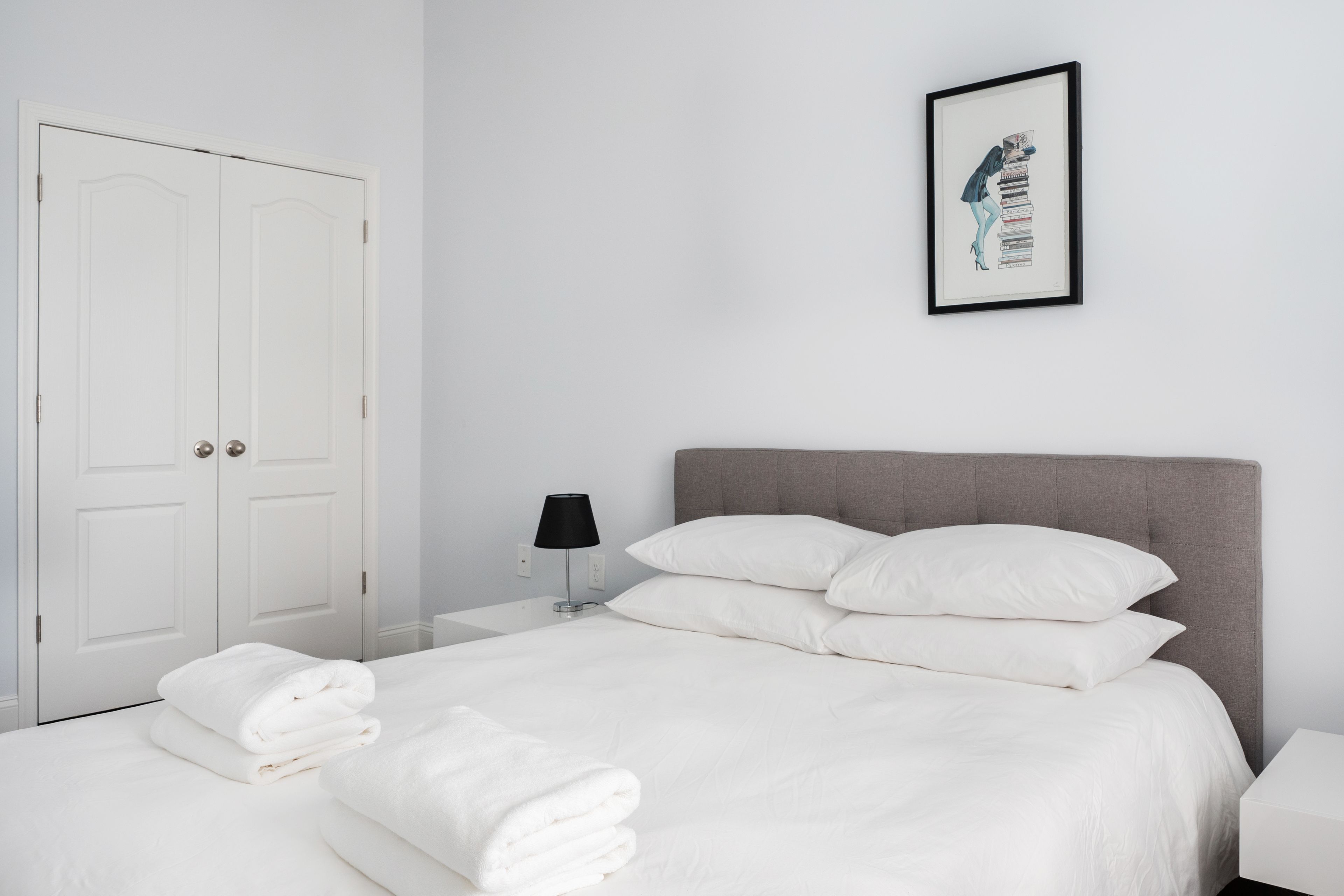 Una cama bien tendida con sábanas blancas y dos almohadas, una mesita de noche con una lámpara y un cuadro en la pared.