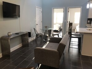 Uma sala de estar com sofá, poltronas, mesa de vidro, TV fixada na parede e porta que dá para outro cômodo.