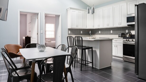 A modern kitchen with white cabinets, a central island, and a dining area with chairs. - Giles Ave 6 Bedroom Gem (Jersey City)