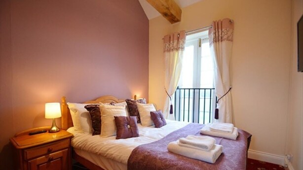 A neatly made bed with a purple bedspread and several pillows, a bedside lamp, and a window with curtains. - The Old Red Lion (Thame)