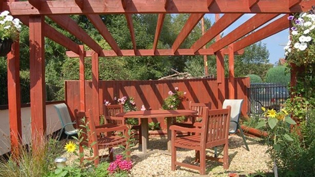 A wooden pergola structure with seating underneath, surrounded by plants and flowers. - The Old Red Lion (Thame)