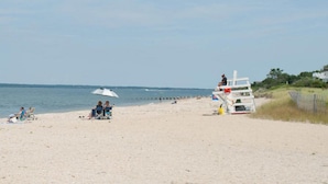 Ein Strand mit einem Rettungsturm, einigen Personen, die auf Stühlen sitzen, und einem weißen Sonnenschirm.